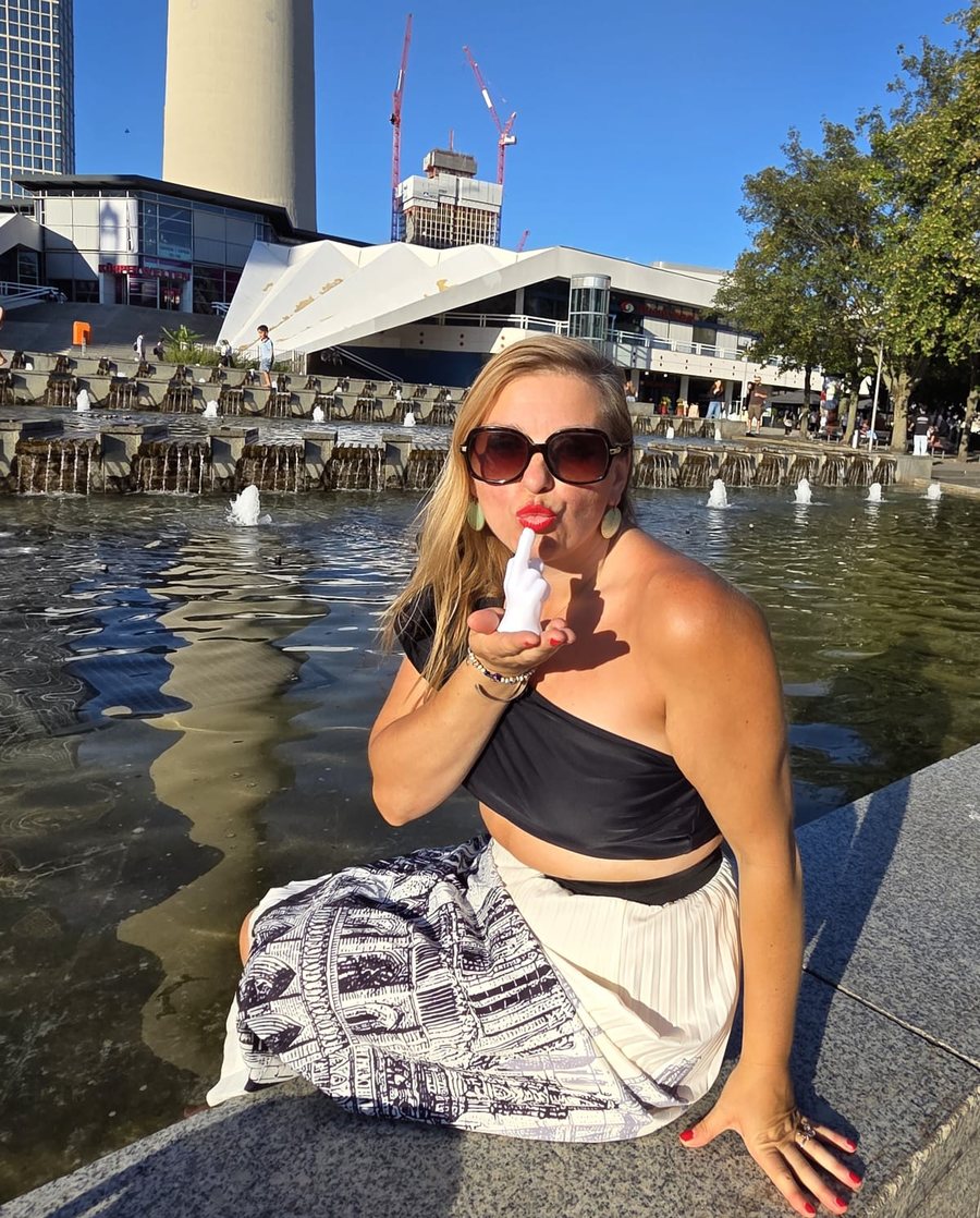 WowMoman sitzt am Brunnen auf dem Berliner Alexanderplatz, Sonnenbrille, roter Lippenstift, und grinst gut gelaunt in die Kamera